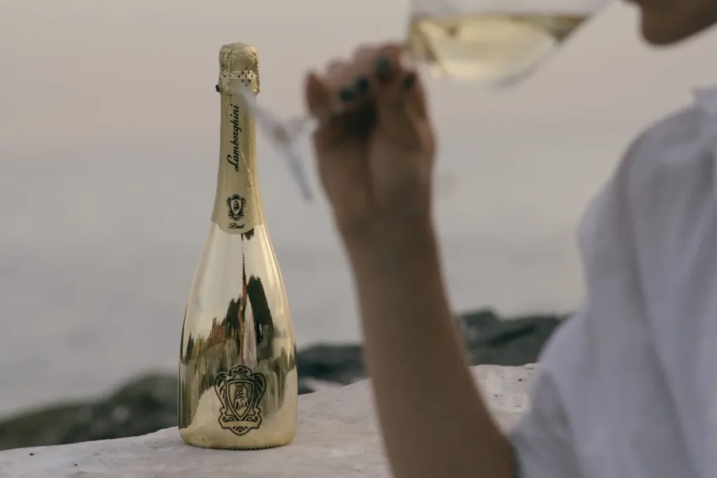 Lamborghini prosecco bottle with a seaside view, while a person in the background enjoys a glass of sparkling wine.