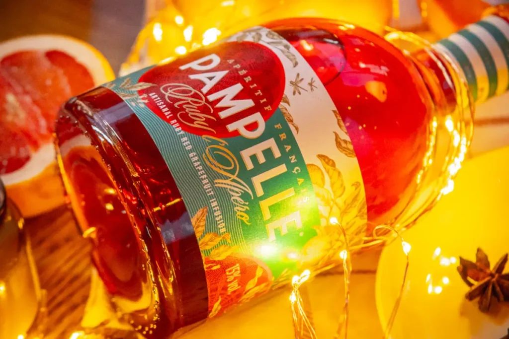 A bottle of Pampelle aperitif surrounded by fresh citrus fruits and decorative lights, highlighting its deep ruby color.