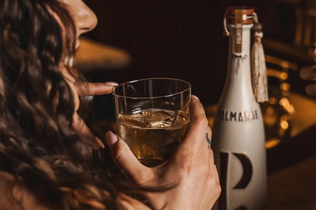 Zbliżenie na osobę trzymającą drink z ginem PALMARÁE z lodem, obok butelki.