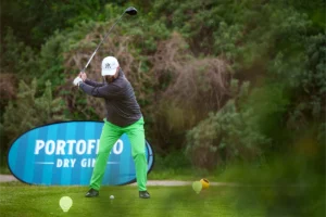 Gracz wykonujący uderzenie na polu golfowym podczas turnieju Golf Business League, w tle niebieski banner sponsora Portofino Dry Gin.