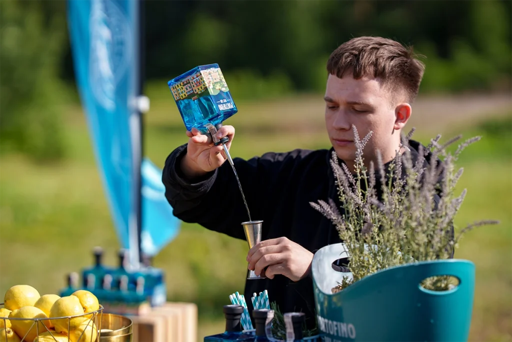Barman w plenerze nalewający rzemieślniczy włoski Portofino Dry Gin z kwadratowej, niebieskiej butelki do miarki barmańskiej na tle pola golfowego.