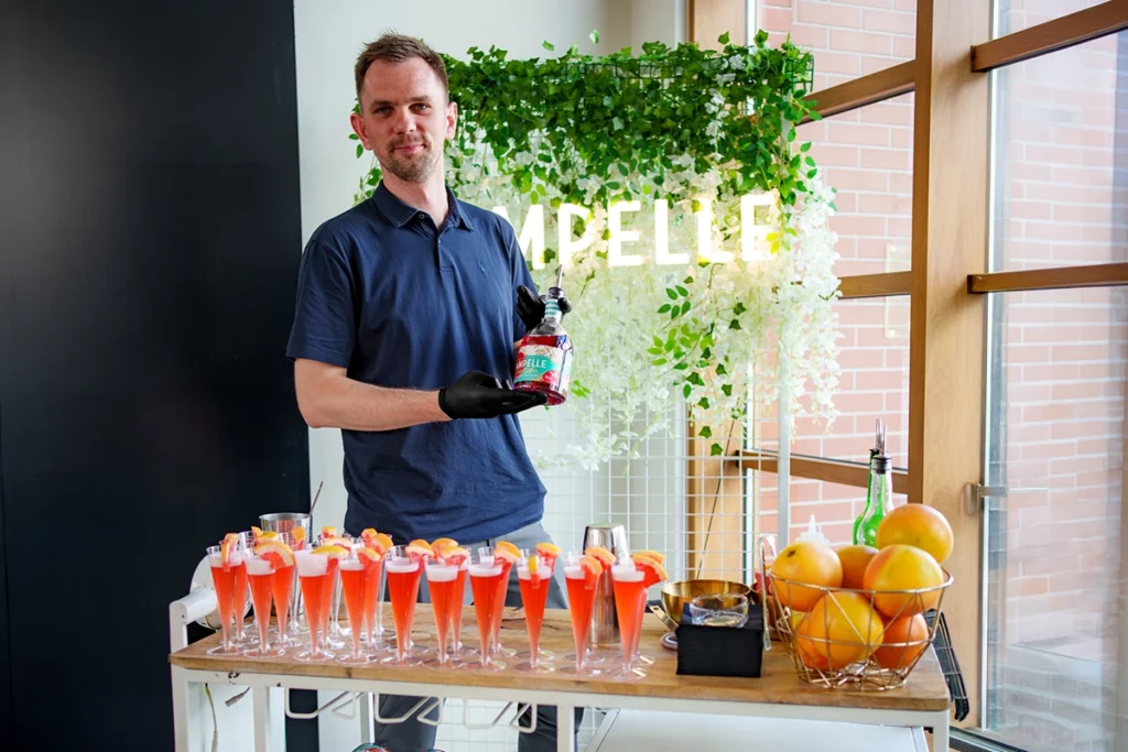 Uśmiechnięty barman prezentujący butelkę francuskiego aperitifu Pampelle przy białym wózku barowym, na którym stoją gotowe, rubinowe koktajle.