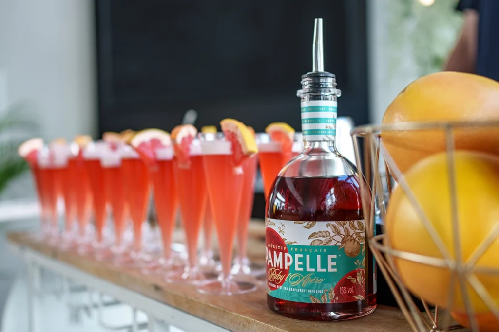 Rząd gotowych, rubinowych koktajli z cząstką grejpfruta w wysokich kieliszkach oraz butelka francuskiego aperitifu Pampelle na drewnianym stole
